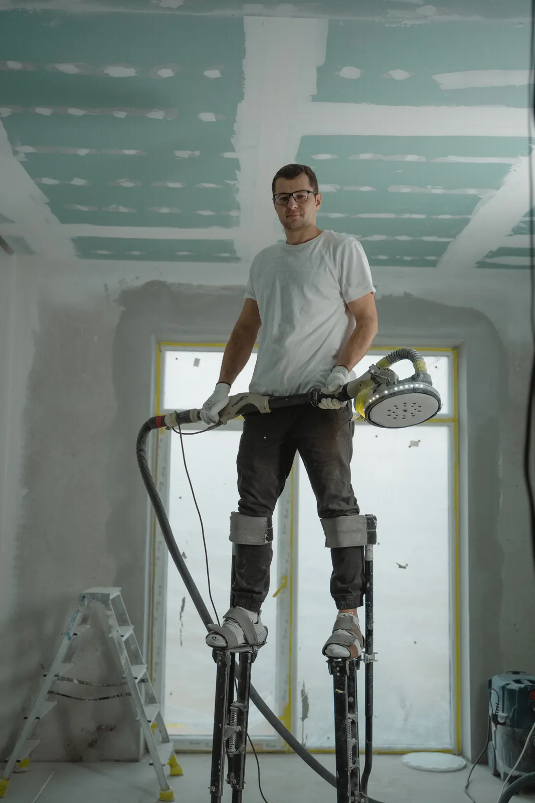 Worker on stilts preparing an unfinished drywall ceiling inside a renovation project.