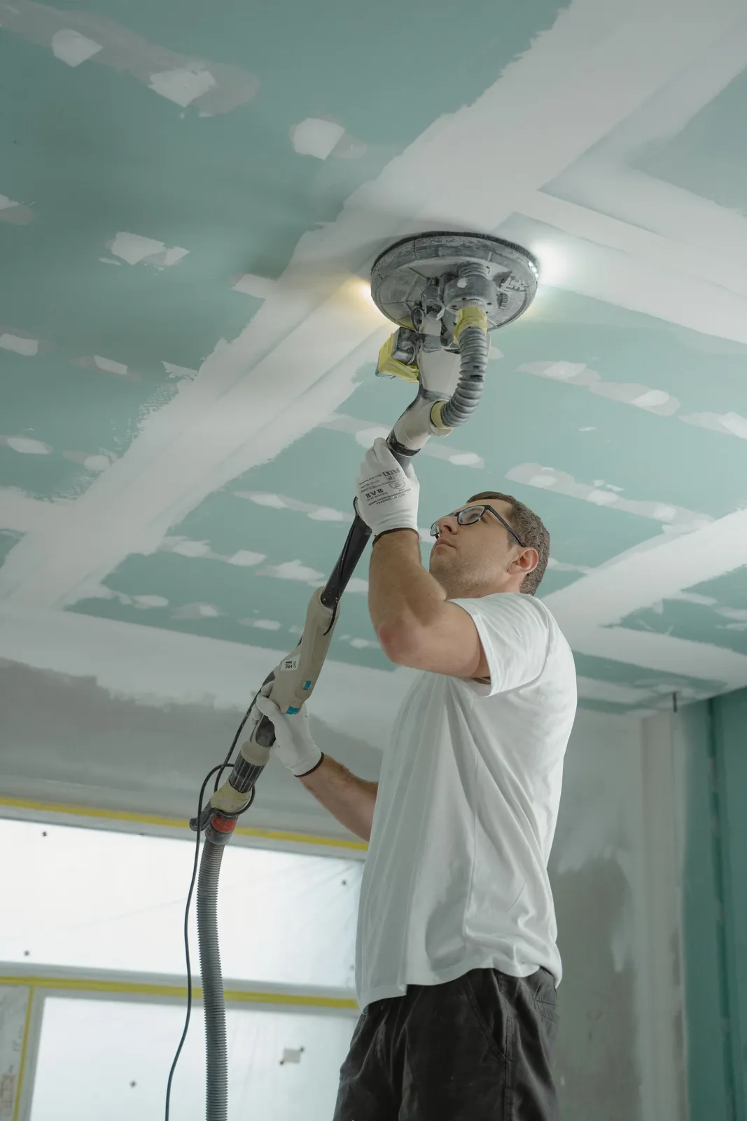 Worker sanding a drywall ceiling with powered dust-control equipment.