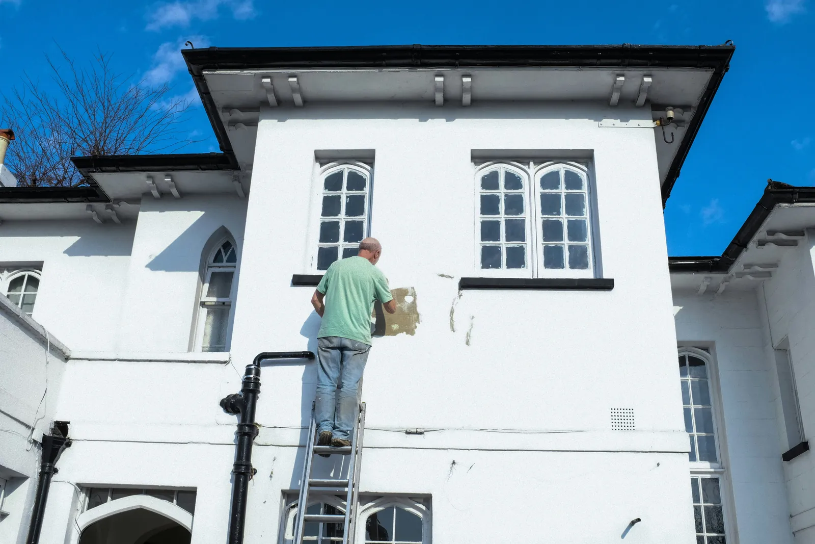 Painter working from a ladder on the white exterior wall of a multi-story home.