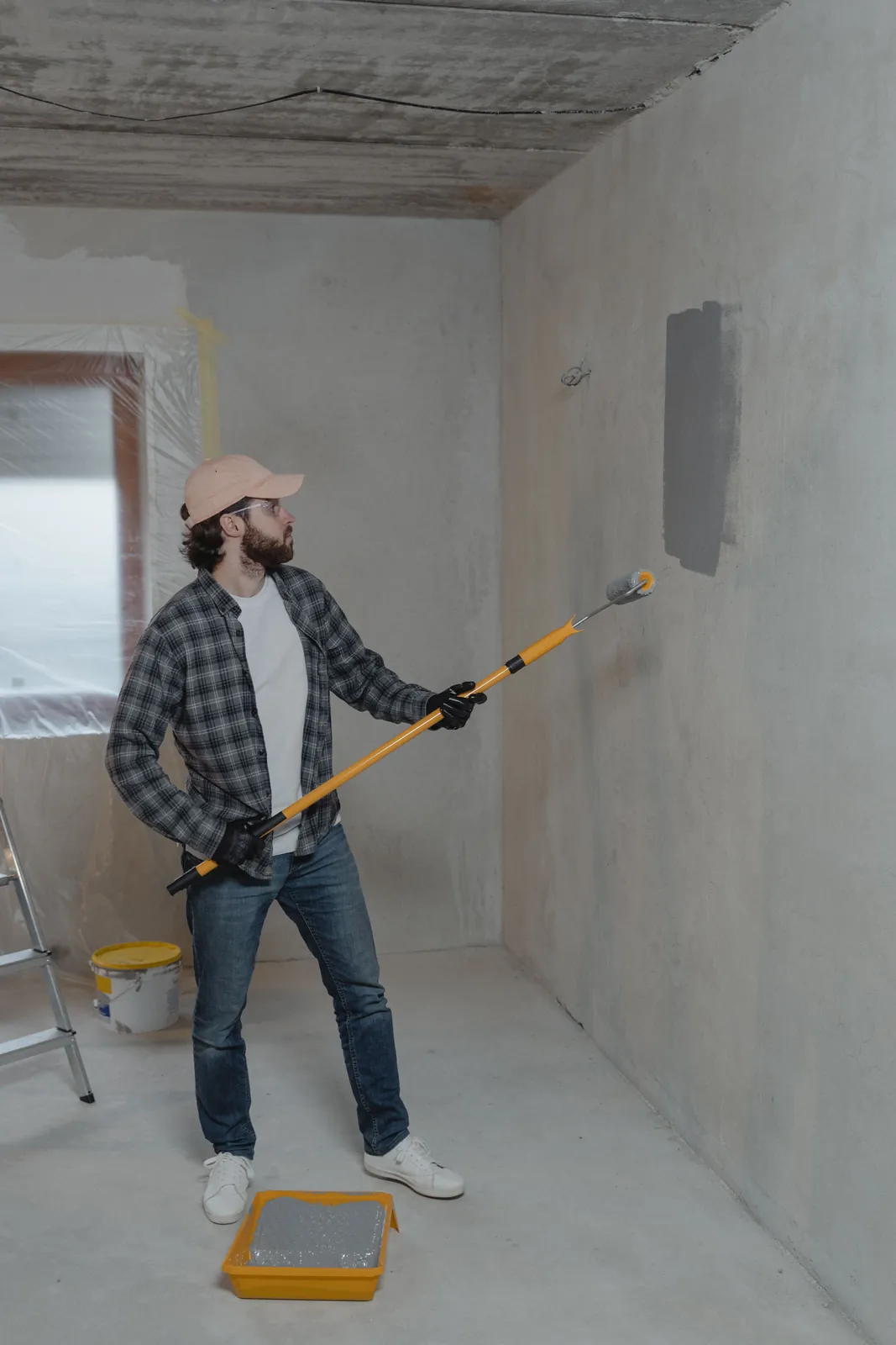 Painter applying gray paint to an unfinished interior wall with a long roller.